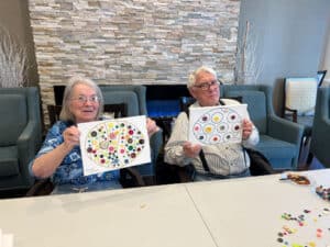 Two elderly people sit at a table in a lounge, proudly showing round boards covered with colorful buttons as artwork.