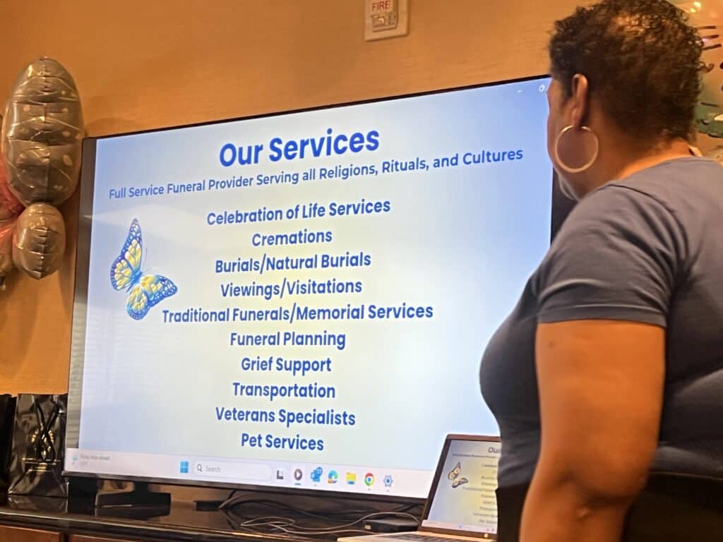 Person in foreground looking at a large screen displaying a slide titled 'Our Services' with funeral-related offerings.",